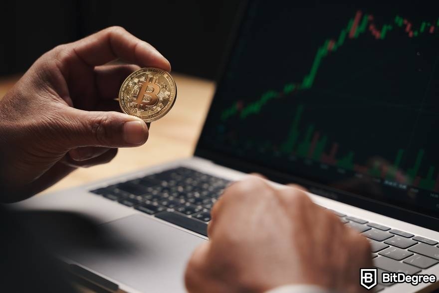 Best-paying jobs in finance: a man holding a physical Bitcoin in front on a laptop.