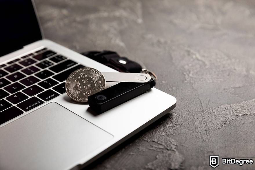 Cara trading crypto: Wallet Ledger dan Bitcoin fisik disimpan diatas sebubah laptop.