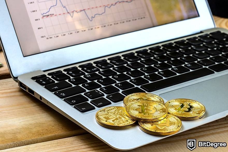 O que é moeda fiduciária em cripto: moedas físicas de Bitcoin na frente de um laptop que mostra dados de mercado.