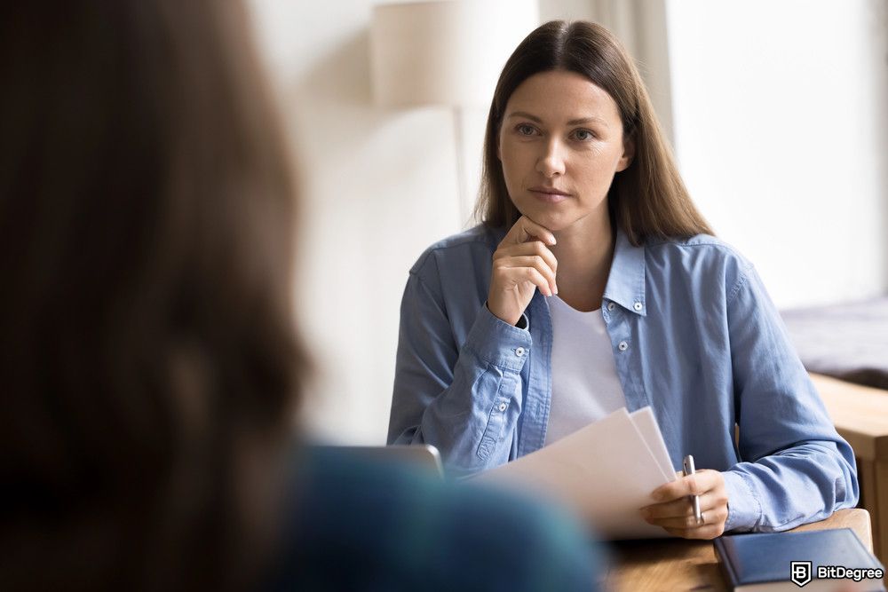 Best product management courses: a serious female HR manager hold resume seated at desk opposite the candidate.