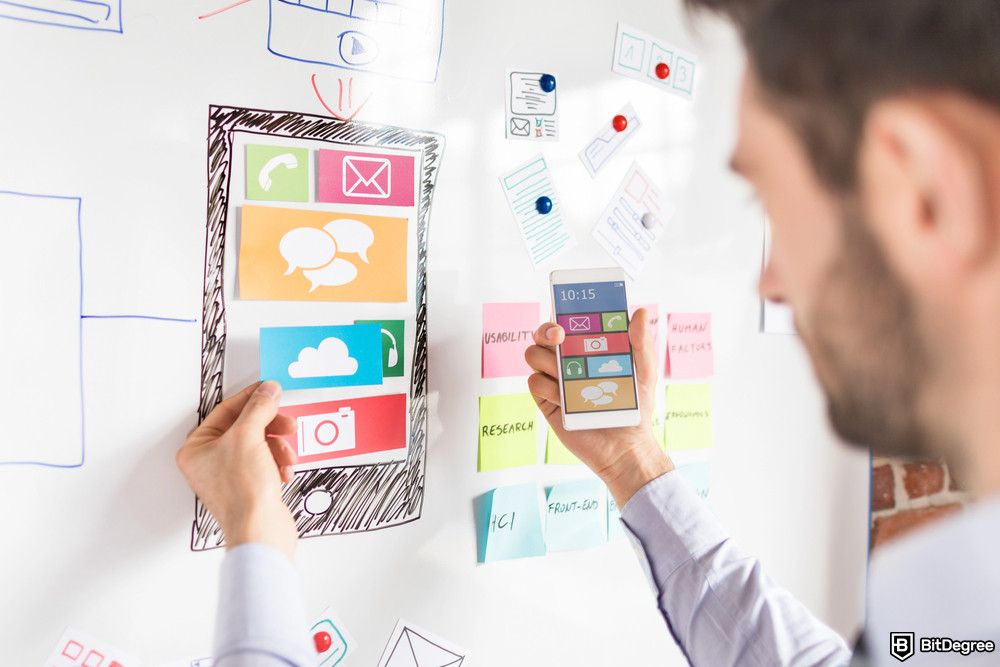 Best product management courses: A man holds a smartphone in their right hand and colorful paper cutouts representing app icons in their left hand.