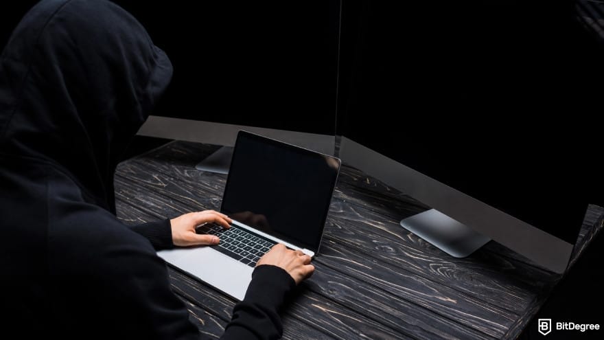 Anonymous casino: hooded person using a laptop near computer monitors. Anonymous casino: hooded person using a laptop near computer monitors.