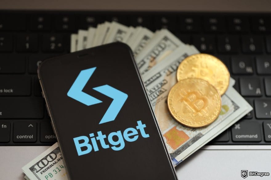 Bitget review: a phone with Bitget logo on a stack of $100 bills and two BTC coins.