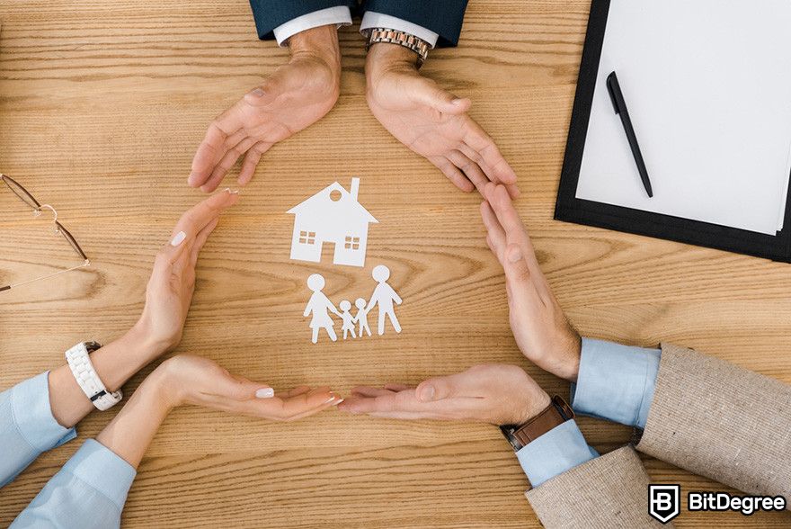 Blockchain in insurance: four hands encircle paper cutouts of a house and family on a wooden table.