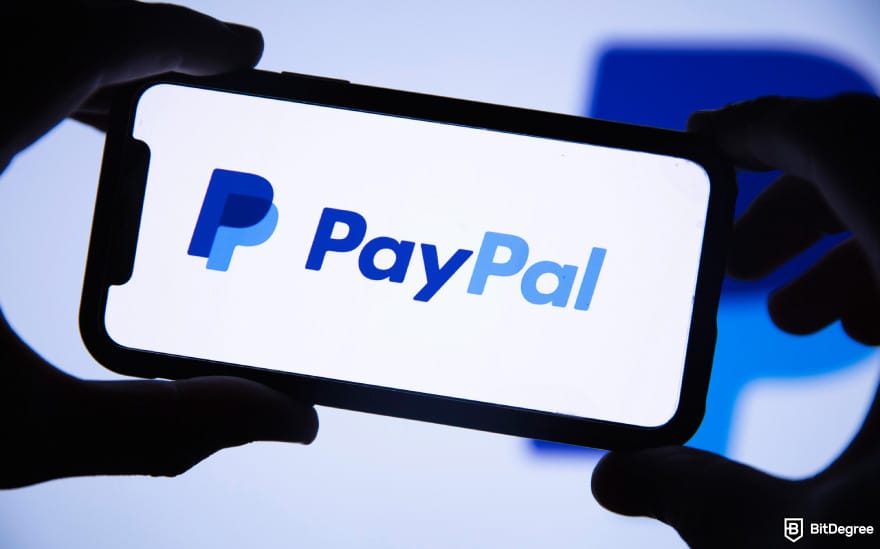 Buy XRP with PayPal: two hands holding a phone with its screen showing PayPal logo.