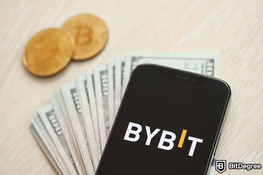 Bybit affiliate: a phone displaying Bybit's logo on top of a stack of 100 USD bills and two BTC coins.