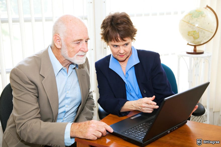 Can you buy crypto on Schwab: a female asset manager showing her laptop screen to an old male client.