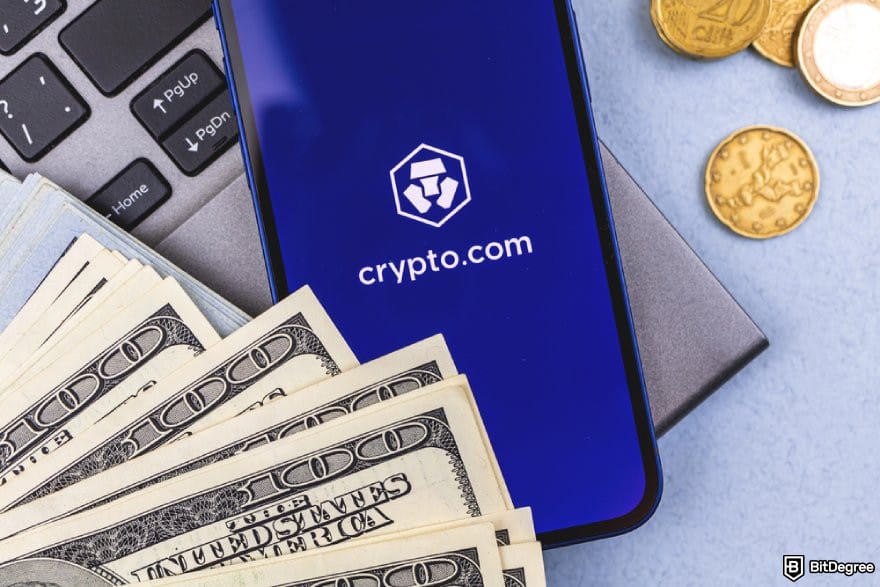Crypto.com review: a smartphone displaying the Crypto.com logo, with dollar bills and coins on a laptop keyboard.