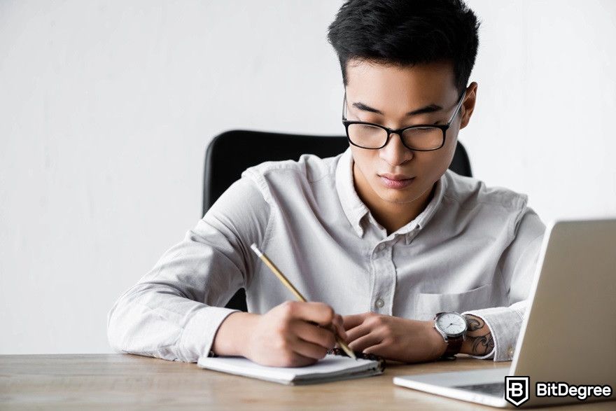 Crypto forums: a man with glasses writing something on a notebook near a laptop.