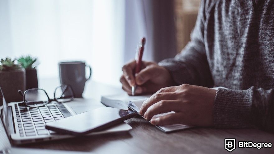 Crypto signals Telegram: Person writing in a book with a laptop, glasses and a cup nearby.