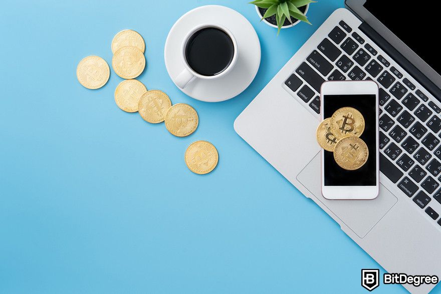 Crypto vs stocks: gold cryptocurrency coins next to a laptop, smartphone, and coffee cup on a blue background.