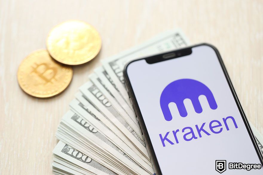 Crypto vs stocks: a mobile phone displaying Kraken logo on top of a stack of 100 dollar bills and two Bitcoin tokens.