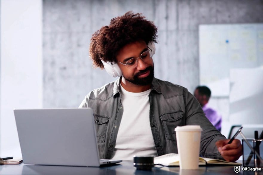 Google Analytics 4 courses: a person wearing a pair of headphones writing on a notebook in front of a laptop.