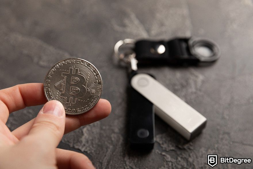 Hardware token: a hand holding a Bitcoin token with a hardware wallet in the background.