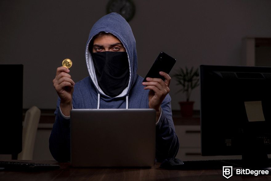 Hardware token: a person wearing a hoodie and a face mask holding a Bitcoin token and a mobile phone in front of a laptop.