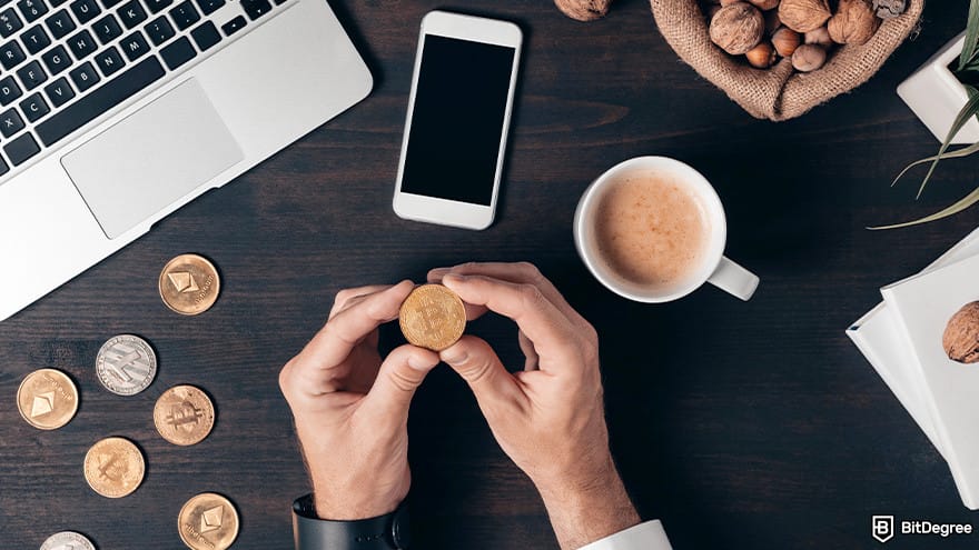 How to cash out Bitcoin: A person holding a Bitcoinwith a coffee cup, a smartphone and a laptop placed on a desk.