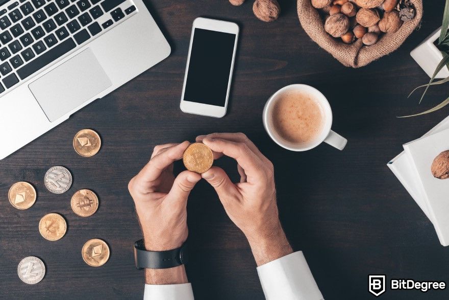 How to cash out Bitcoin: A person holding a Bitcoinwith a coffee cup, a smartphone and a laptop placed on a desk.