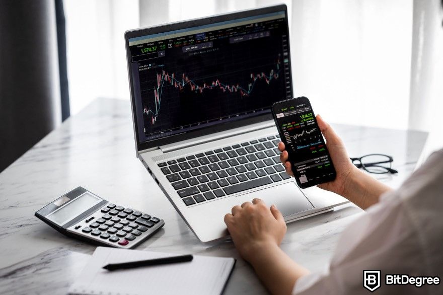 How to cash out Bitcoin: A person looking at a trading dashboard on a smartphone and a laptop with a calculator on the desk.