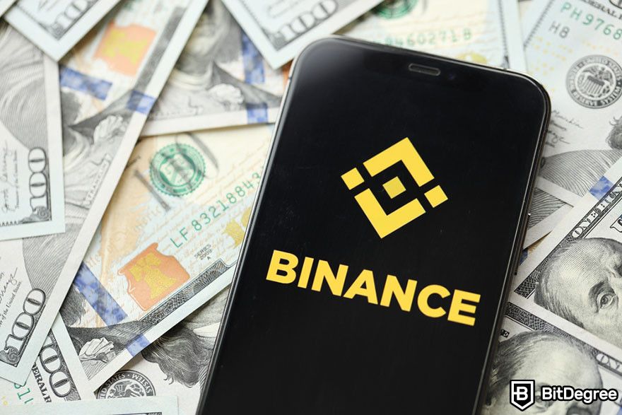 How to get a crypto wallet: A phone with Binance logo on top of a pile of hundred dollar bills.