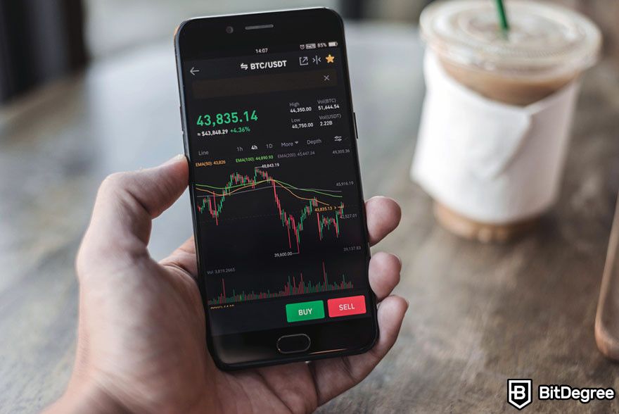 How to read candlesticks: a phone displaying a crypto trading app, held over a cafe table.