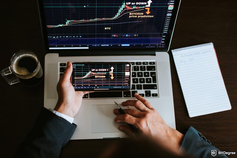 How to short on Coinbase: overhead shot of a person's hands holding a smartphone displaying a cryptocurrency chart, with a similar chart visible on a laptop screen behind it.