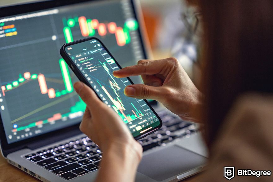 How to short crypto: Close-up of a person analyzing stock market charts on a smartphone with a laptop in the background.