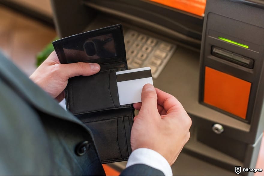 How to withdraw money from Wisely card: a person taking their card out of the wallet in front of an ATM. How to withdraw money from Wisely card: a person taking their card out of the wallet in front of an ATM.