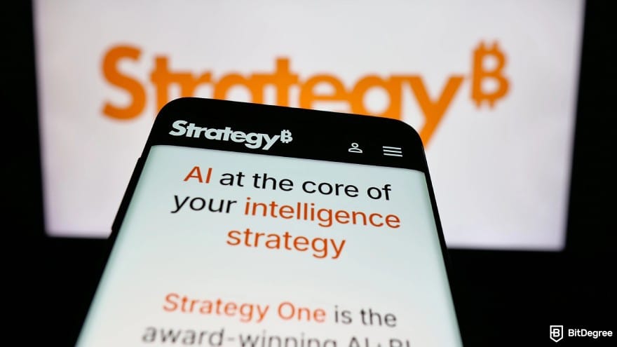 Is Cardano dead: a smartphone screen showing MicroStrategy website in front of a monitor with StrategyB written in the backdrop.