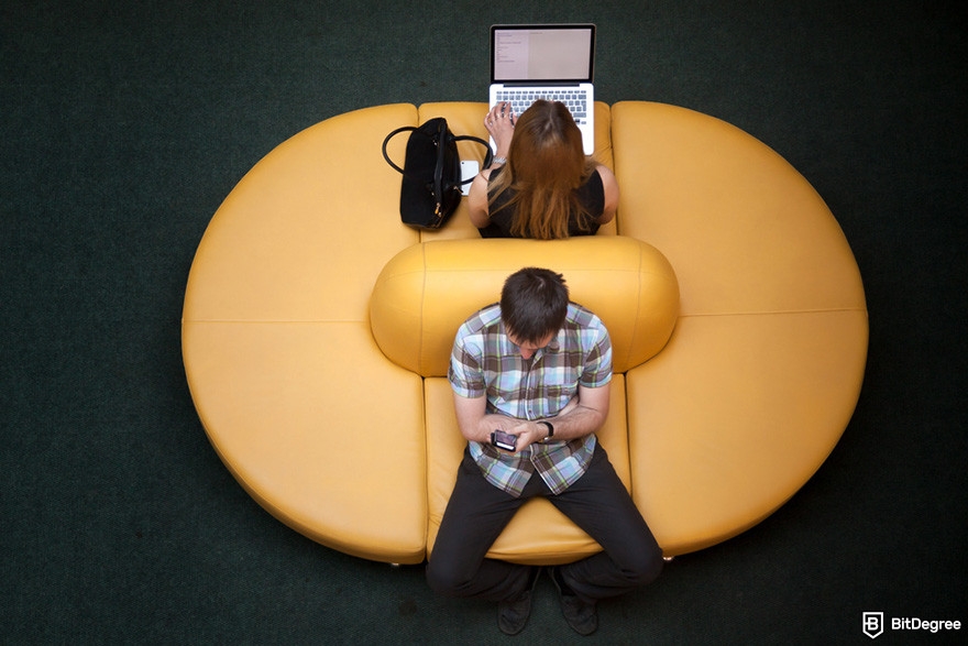 Is MetaMask safe: two people back-to-back on a yellow sofa, one using a laptop, the other a smartphone.