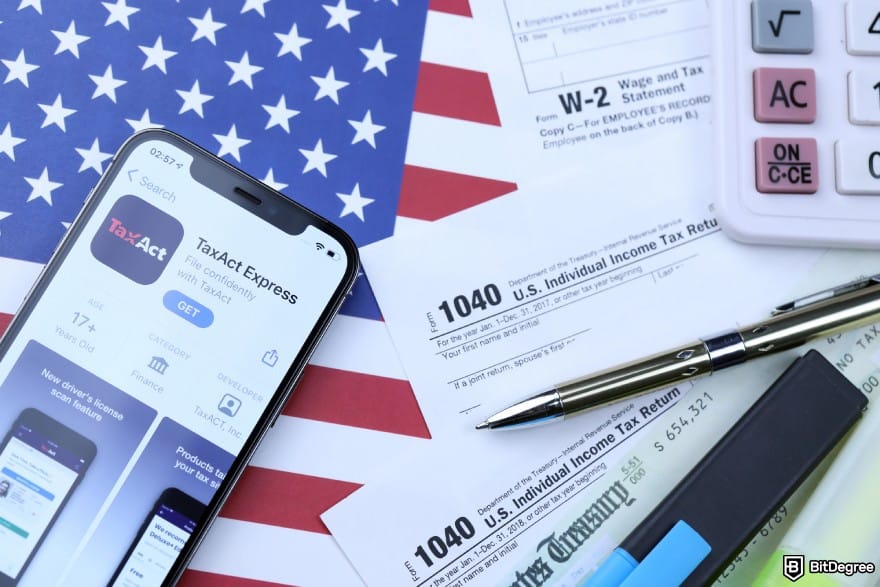 Ledger taxes: a mobile phone showing the TaxAct app on top of an American flag next to tax filing forms, a calculator, and stationery.
