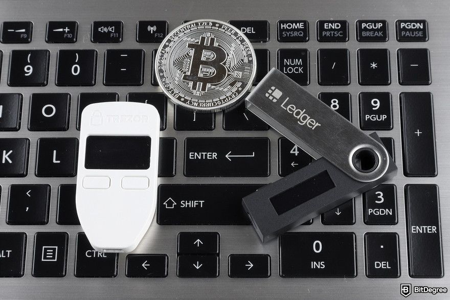 Managing multiple crypto wallets: a close-up of Trezor wallet, Ledger wallet, and a Bitcoin physical coin on a laptop keyboard.