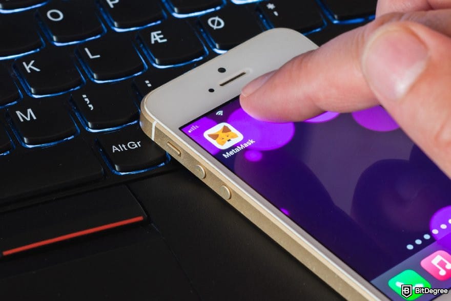 MetaMask review: a finger taps the MetaMask app icon on a white smartphone.
