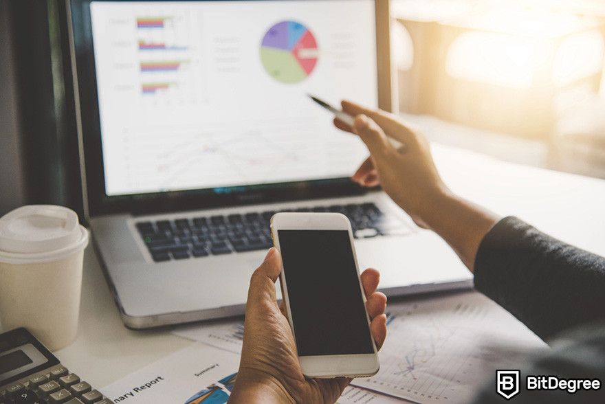 NFT marketing agency: a person holding a mobile phone while pointing at a laptop screen showing charts.