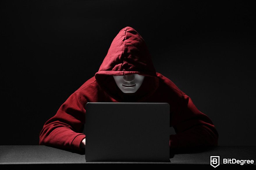 Should I buy Bitcoin: A person wearing a red hoodie and a mask sitting in front of a laptop.