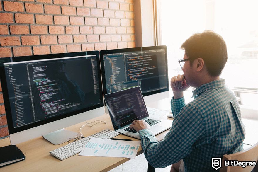 Web3 jobs: a person in a checked shirt works on a laptop with code on the screen, surrounded by multiple monitors displaying code in a modern office.