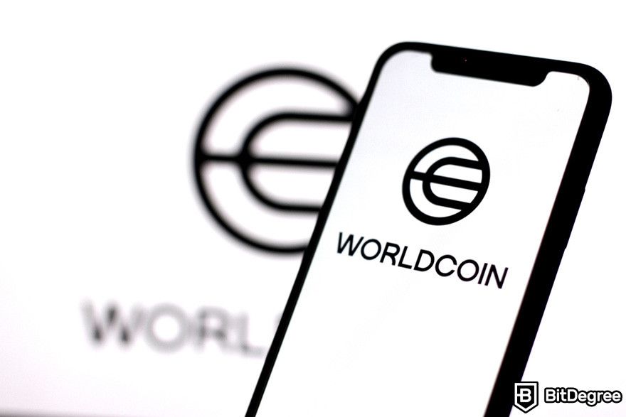 Worldcoin là gì: một chiếc điện thoại di động hiển thị logo của Worldcoin ở phía trước nền trắng với một logo Worldcoin khác.