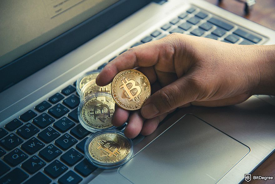 Withdrawal Bitstamp: a person holding physical Bitcoin coins above a laptop keyboard.