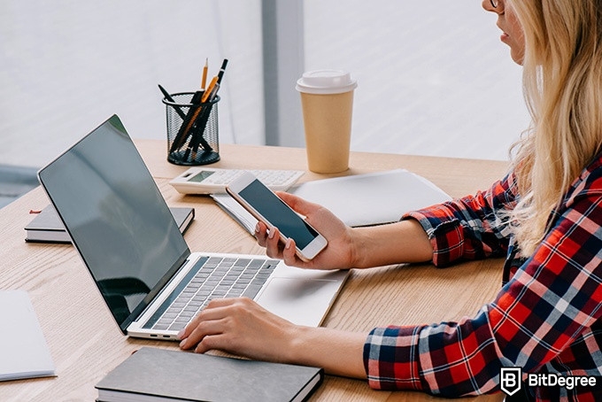 Online information technology degree: a woman is holding a phone and looking at a laptop.