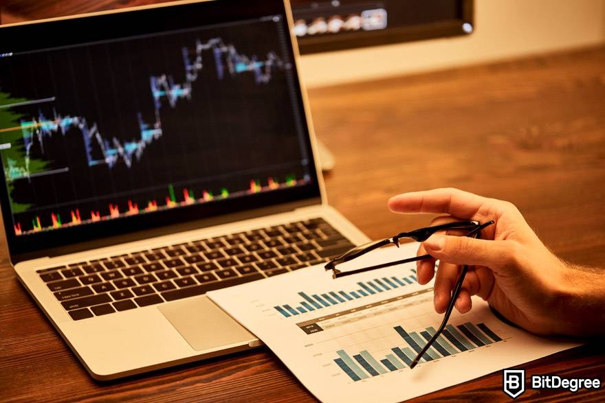 Bet crypto trading course: a hand is holding glasses and pointing at a market chart on a laptop.