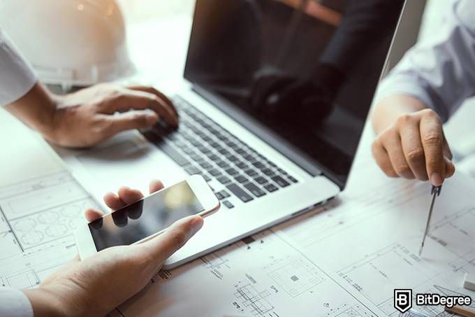 Engineering online degree: one man is holding a phone and using a laptop while another man is checking blueprints.